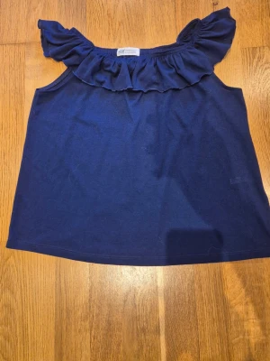 Marinblå volangtopp från H&M - Supersöt marinblå topp från H&M med volangdetalj längs halsringningen. Ärmlös modell perfekt för varma dagar. Enkel att matcha med både jeans och kjol.