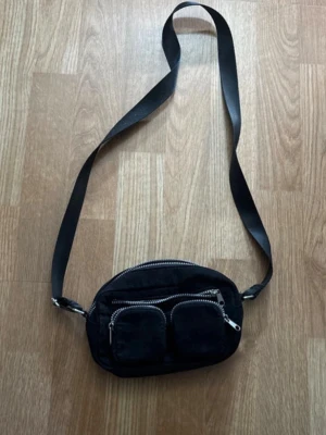 Svart axelväska med flera fack - Snygg svart axelväska i tyg med justerbart band och tre dragkedjeförsedda fack framtill. Perfekt för att hålla ordning på småsaker. Väskan har en stilren och enkel design med silvriga detaljer.
