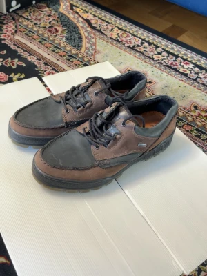 Bruna och svarta loafers från Ecco - Säljer ett par snygga loafers från Ecco i brunt och svart läder. Skorna har robust sula, svarta snören och klassisk seglarsko-design. Perfekta för dig som gillar stilrena och bekväma skor med sportig känsla.