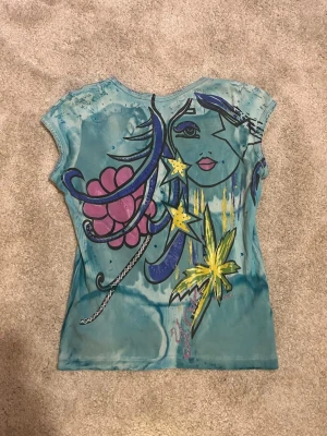 Y2k tie-dye topp från Yoki - Unik blå tie-dye topp från Yoki med färgglad grafisk print på ryggen som föreställer ett ansikte, stjärnor och blommor. Toppen har korta ärmar och snörning där fram. Perfekt för dig som gillar statement-plagg och konstnärliga detaljer. Kan se att en liten paljett har försvunnit men det är inget man tänker på. Skulle säga att den passar en XS också. Är gjord 100% av bomull