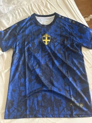 Blågult fotbollströja Adidas - Säljer en blå och svart mönstrad fotbollströja från Adidas med Sveriges landslagsemblem på bröstet. Tröjan har korta ärmar, rund hals och detaljer i gult. Materialet är lätt och andas, perfekt för träning eller match. Limited edition 'Blågult' 2026.