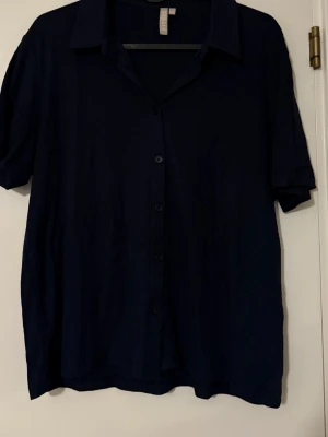 Mörkblå skjorta från Pieces - Stilren mörkblå skjorta från Pieces med klassisk krage och korta ärmar. Skjortan har knäppning framtill och en avslappnad passform, perfekt för en trendig och enkel look.