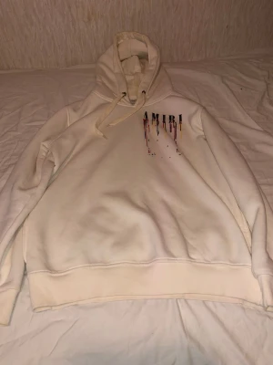 Vit hoodie från Amiri med färgstänk - Vit hoodie från Amiri med huva och dragsko. Tryck med Amiri-logga och färgstänk i rött, gult, blått och svart både fram och bak. Oversized passform och mjukt material, perfekt för en trendig streetwear-look.