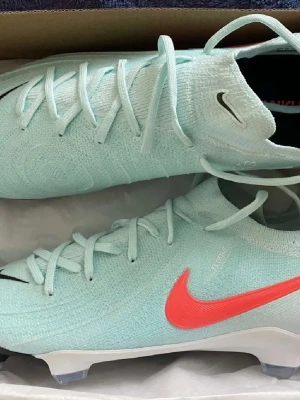 Nike Mercurial ljusblå fotbollsskor - Snygga Nike Mercurial fotbollsskor i ljusblått med en röd Swoosh-logga på sidan. Skorna har stickad ovandel för bra passform och känsla, samt fasta dobbar för spel på gräs. Lätta och smidiga med modern design, perfekta för snabba spelare som vill sticka ut på planen.