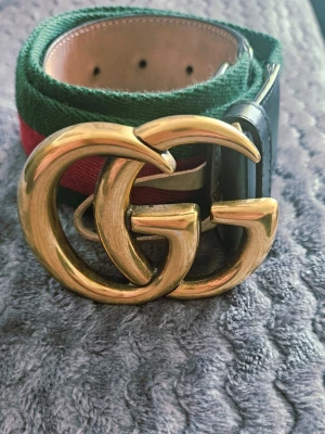 Gucci bälte med GG-spänne 80/32 - Snyggt Gucci-bälte med ikoniskt dubbelt GG-spänne i guld. Bältet har klassiska gröna och röda ränder samt detaljer i svart och beige skinn. Perfekt accessoar för att lyfta vilken outfit som helst. Köparen står för frakt ej retur. 