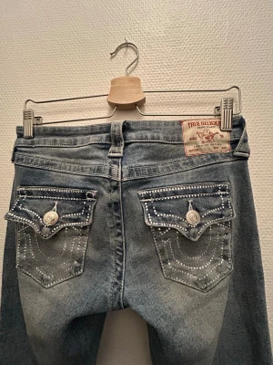 True Religion ljusblå bootcut jeans med glitter - Säljer ett par ljusblå jeans från True Religion med bootcut-modell. Jeansen har dekorativa fickor bak med vita sömmar och silverfärgade knappar. Klassisk patch på linningen och snygg tvättad look. Perfekta för dig som gillar detaljer och streetstyle. sytt upp dom så det passar mig (jag är ungefär 165 cm) skriv om du är intresserad💞använd någon enstaka gång! säljer pga kommer inte till användning.