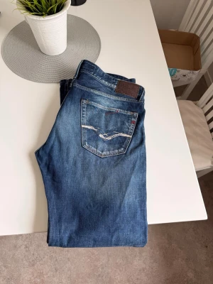 Replay jeans  - Säljer ett par blå jeans från Replay med snygga slitningar och kontrastsömmar på bakfickan. Klassisk femficksmodell med normal passform och läderpatch bak i midjan. Perfekta till en avslappnad stil och tillverkade i mjukt denimtyg.
