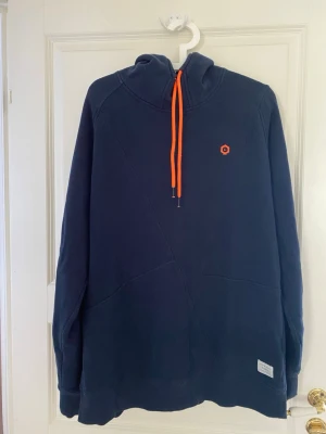 Mörkblå hoodie med orange snören - Snygg mörkblå hoodie från WeSC med huva och orange dragsko. Liten orange logga på bröstet och en diskret etikett nertill. Mjuk bomullskänsla och avslappnad passform, perfekt för chill dagar.