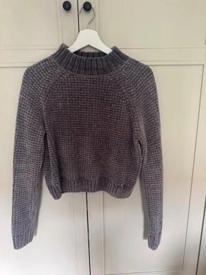Grå stickad tröja från H&M Divided - Mysig grå stickad tröja från H&M Divided med grov ribbstickad krage, muddar och nederkant. Tröjan har en avslappnad passform och är tillverkad i ett mjukt, chenille-liknande material som känns skönt mot huden.