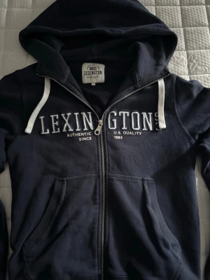 Marinblå hoodie från Lexington - Snygg marinblå hoodie från Lexington med dragkedja och vita snören i huvan. Stor Lexington-logga broderad över bröstet och två rymliga fickor framtill. Tillverkad i mjuk bomullsblandning som känns skön mot huden. Perfekt för en avslappnad och sportig stil.