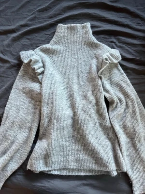 Ljusgrå stickad polotröja med volang - Mysig ljusgrå stickad polotröja med volangdetaljer på axlarna. Tröjan har lång ärm och är tillverkad i ett mjukt, fluffigt material som känns skönt mot huden. Perfekt för lager-på-lager och en trendig look.