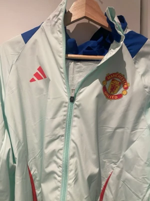 Manchester United träningsjacka Adidas - Ljusblå träningsjacka från Adidas med Manchester Uniteds klubbmärke på bröstet. Jackan har hel dragkedja, huva, blå detaljer på axlarna och röda inslag vid fickorna. Perfekt för fotbollsträning eller som sportig streetwear.