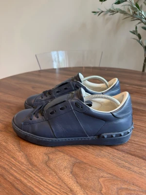Mörkblå sneakers från Valentino - Snygga mörkblå sneakers från Valentino i läder med svarta skosnören och ikoniska nitar på sulan. Skorna har en stilren design med låg profil och ljus insida. Perfekta för dig som gillar exklusiva och trendiga märken. Passar även 42
