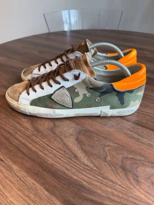 Camo sneakers från Philippe Model - Snygga sneakers från Philippe Model med camouflagemönster, bruna mockadetaljer och orange hälparti. Skorna har vita och gröna partier i textil, bruna skosnören och låg profil. Perfekta för dig som vill sticka ut med en unik stil.