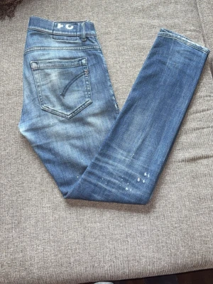 Dondup jeans - Säljer ett par blå jeans med snygga slitningar och distressed detaljer framtill. Jeansen har smal passform, klassiska fem fickor och coola färgstänk. Perfekta för en avslappnad streetstyle-look.