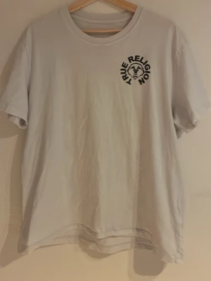 Vit True Religion t-shirt med tryck - Vit t-shirt från True Religion med svart tryck på bröstet och stort grafiskt tryck med text på ryggen. Klassisk rund hals och korta ärmar. Perfekt för en avslappnad och trendig stil.