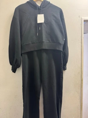 Svart set från Puma med huva - Snyggt svart set från Puma med croppad hoodie och matchande byxor. Hoodien har broderad Puma-logga framtill och dragsko i huvan. Byxorna har slits vid benslut och är i mjukt, bekvämt material. Perfekt för en avslappnad och sportig look.