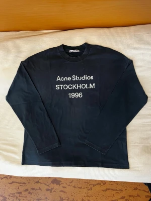 Långärmad Acne Studios tröja - Svart långärmad t-shirt från Acne Studios med vit text 'Acne Studios STOCKHOLM 1996' på bröstet. Klassisk rund hals och rak passform. Perfekt för en stilren och avslappnad look. 59cm bröst 70cm längd. Fraktar inom 24h! Nästan oanvänd, bara testad!