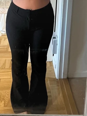 Svarta utsvängda kostymbyxor - Snygga svarta kostymbyxor med utsvängda ben och hög midja. Klassisk design med fickor bak och knappstängning fram. Perfekta för en stilren look och passar till många olika outfits.