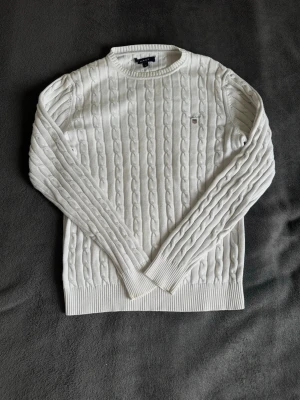 Vit kabelstickad tröja från Gant - Snygg vit kabelstickad tröja från Gant med rund halsringning och broderad logotyp på bröstet. Tröjan har långa ärmar och ribbade muddar vid ärmslut och nederkant. Perfekt för en stilren och klassisk look.