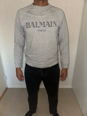 Grå sweatshirt från Balmain Paris - Snygg grå sweatshirt från Balmain Paris med svart logotyptryck på bröstet. Tröjan har rund halsringning, långa ärmar och är tillverkad i mjukt material som passar perfekt till en avslappnad stil.
