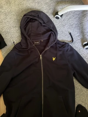 Svart hoodie från Lyle & Scott - Svart hoodie med dragkedja från Lyle & Scott. Klassisk design med huva och snörning, samt gul broderad logga på bröstet. Perfekt för en avslappnad och stilren look. Mjukt material och fickor framtill.