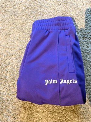 Lila mjukisbyxor från Palm Angels - Säljer ett par lila mjukisbyxor från Palm Angels med vit logga på benet och vita sidoränder. Byxorna har resår i midjan och raka ben. Perfekta för en chill och trendig look.
