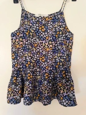 Blommig linnetopp från H&M - Supersöt linnetopp från H&M med blommigt mönster i lila, gul, rosa och vitt på mörk botten. Tunt tyg med smala axelband och volang nertill. Perfekt för en färgglad och chill look.