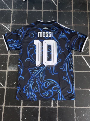 Argentina Messi matchtröja blå Adidas - Snygg Argentina matchtröja från Adidas med Messi och nummer 10 på ryggen. Tröjan är svart med blåa och ljusblåa mönster, vita detaljer och emblem på bröstet. Tillverkad i lätt och ventilerande material, perfekt för fotboll eller träning.