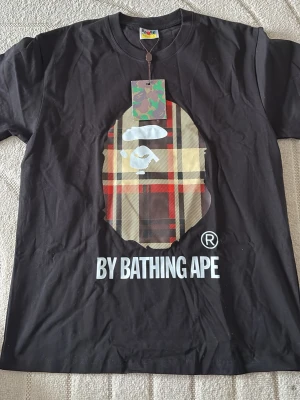 Svart BAPE t-shirt med rutig logga - Säljer en svart t-shirt från BAPE med den ikoniska apansiluetten i rutig design på bröstet och texten 'BY BATHING APE' under. T-shirten har rund hals och korta ärmar. Materialet är mjuk bomull och den har en streetwear vibe. Sitter som M!