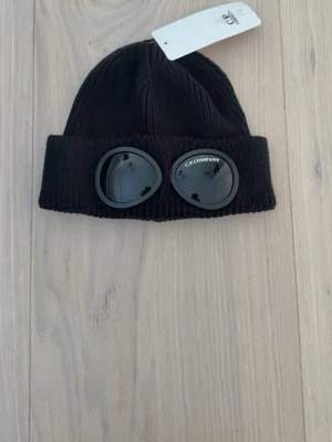 Svart mössa med goggles C.P. Company - Cool svart ribbstickad mössa från C.P. Company med ikoniska goggles framtill. Mössan har en uppvikt kant och är tillverkad i ett mjukt stickat material. Perfekt accessoar för att lyfta din streetwear-stil.