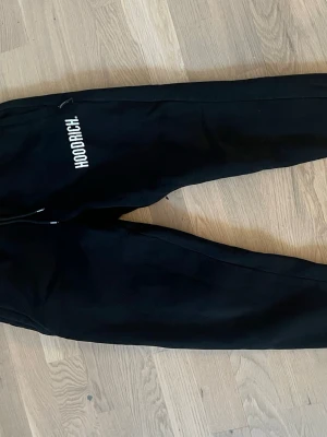 Hoodrich byxor - Svarta mjukisbyxor från Hoodrich med vit logga på ena benet. Byxorna har dragsko i midjan och är tillverkade i mjukt material med avslappnad passform. Perfekta för en chill och street-inspirerad stil.
