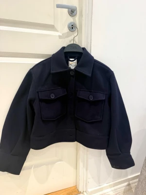 Mörkblå cropped cardiganjacka Zara - Snygg mörkblå cardiganjacka från Gina Tricot med två stora bröstfickor och knappar framtill. Jackan har en rak, boxig passform och klassisk krage. Perfekt för lager-på-lager och ger en cool, trendig look. Materialet är tjockt och känns mjukt. Helt nytt skick, lappen sitter fortfarande kvar. Passar xxs/xs