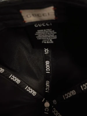 Svart keps från Gucci med GG-mönster - Svart keps från Gucci med klassiskt GG-monogram över hela kepsen och en grön-röd rand på sidan. Kepsen är tillverkad i tyg och har detaljer med Gucci-logga på insidan. Perfekt accessoar för en trendig look.