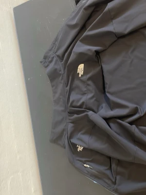 Svart vindjacka från The North Face - Snygg svart vindjacka från The North Face med dragkedja och logga på bröstet. Jackan har praktiska fickor med dragkedja och är tillverkad i ett lätt och slitstarkt material som passar perfekt för blåsiga dagar.