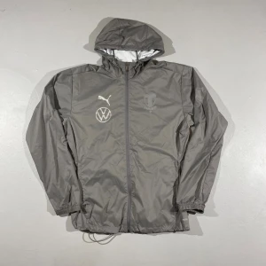 MFF/Puma vindjacka - Snygg grå windbreaker från Puma/ Malmö FF. VW-märke på bröstet. Jackan har huva, hel dragkedja och elastiska muddar. Perfekt för regn blåst och sportiga outfits.