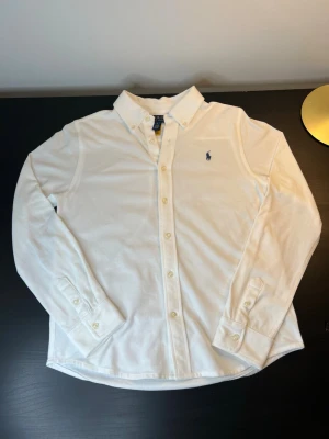 Vit skjorta från Polo Ralph Lauren - Klassisk vit skjorta från Polo Ralph Lauren med broderad blå logga på bröstet. Skjortan har lång ärm, knappar framtill och är tillverkad i bomull. Perfekt för en clean och stilren look.