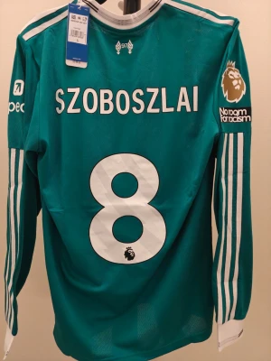 Liverpool spelar Version långärmad matchtröja teal - Snygg tealgrön långärmad Liverpool FC matchtröja från Adidas med vita detaljer och tryck. Tröjan har Szoboszlai och nummer 8 på ryggen, Premier League-märke och 'No room for racism' på ärmen. Tillverkad i lätt och ventilerande material, perfekt för fotbollsträning eller match.
