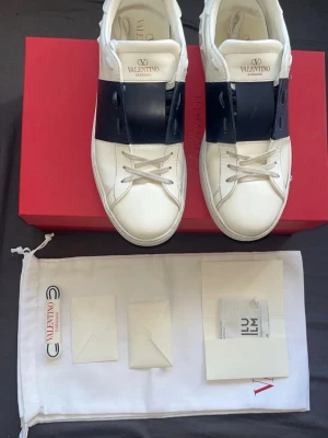 Valentinos open mörkblå - Snygga vita sneakers från Valentino med en bred svart läderdetalj över vristen. Klassisk låg modell med rund tå och vita snören. Märkeslogga på plösen och insidan. Kommer med originalkartong och dustbag.
