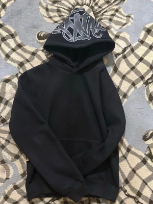 Syna World Hoodie - Svart hoodie med stor huva och unikt graffiti-inspirerat tryck i vitt på huvan. Klassisk passform med ribbade muddar och magficka. Perfekt för dig som gillar streetwear och vill sticka ut med en cool detalj.