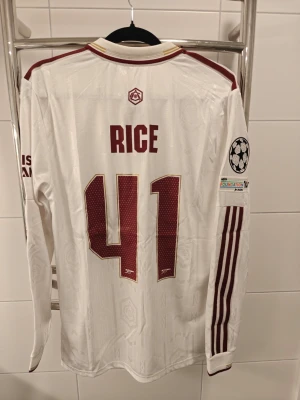 Arsenal långärmad matchtröja Rice 41 spelar version - Spelar version Vit långärmad Arsenal matchtröja från Adidas med vinröda detaljer, spelarnamn Rice och nummer 41 på ryggen. Tröjan har Champions League-märke på ärmen, klassisk krage och broderade loggor. Materialet är lätt och andas, perfekt för fotbollsträning eller match.