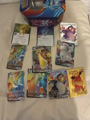 Pokémonkort samling med plåtlåda - Samling av Pokémonkort med olika motiv, inklusive flera holografiska och VMAX-kort samt en guldfärgad Pikachu-kort. Medföljer en färgglad plåtlåda med Pokémonmotiv. Perfekt för samlare eller Pokémonfans som vill utöka sin kortsamling.