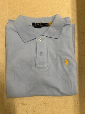 Ralph Lauren piké  - Klassisk ljusblå pikétröja från Ralph Lauren med gul broderad logga på bröstet. Tröjan har krage, knappar framtill och är tillverkad i bomull. Perfekt för en stilren och avslappnad look.
