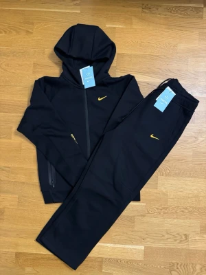 Svart NOCTA x Nike tech fleece set - Svart hoodie och matchande byxa från NOCTA x Nike med gula loggor. Produkten är helt oanvänd, vill sälja för storleken passade inte. Storleken är egentligen L storlek men den passar för M. Priset kan diskuteras.