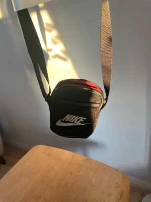 Svart axelväska från Nike - Säljer en svart axelväska från Nike med vit logga på framsidan. Väskan har justerbar axelrem, dragkedja och vinrött innerfoder. Perfekt storlek för det viktigaste och enkel att bära crossbody. Materialet är slitstark textil.