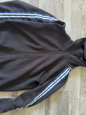 Svart hoodie med blåa ränder - Svart ifk hoodie från kommandobryggan huvan har även maskering. Ops helhet fint skick men några skador i änden på armen. St M sitter som S
