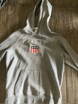 Grå hoodie från GANT med logga - Snygg grå hoodie från GANT med broderad logga och sköld med stjärnor och ränder på bröstet. Klassisk känguruficka framtill och dragsko i huvan. Perfekt för en avslappnad och sportig stil.