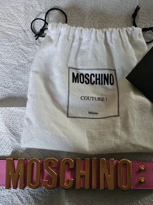 Rosa Moschino armband med guldtext - Säljer ett statementarmband från Moschino i rosa läder med stora guldfärgade bokstäver framtill. Armbandet har bred design och justerbar knäppning med fyra guldfärgade knappar. Perfekt accessoar för att lyfta din outfit.,