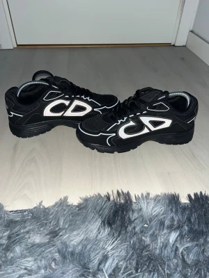 Svarta Dior sneakers med vita detaljer - Säljer ett par svarta Dior sneakers med stora vita CD-loggor på sidorna och coola reflekterande detaljer. Skorna har en chunky sula och meshpartier för extra komfort. Perfekta för dig som vill sticka ut med en lyxig streetstyle-look.