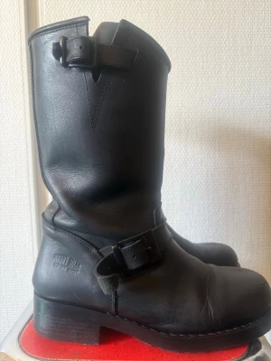 Svarta läderstövlar från Primeboots - Klassiska svarta stövlar i äkta läder från Primeboots med två spännen, ett vid vristen och ett upptill. Grov sula och robust design som ger en cool och tidlös look. Perfekta för dig som gillar bikerstil eller vill ha ett par boots som sticker ut.